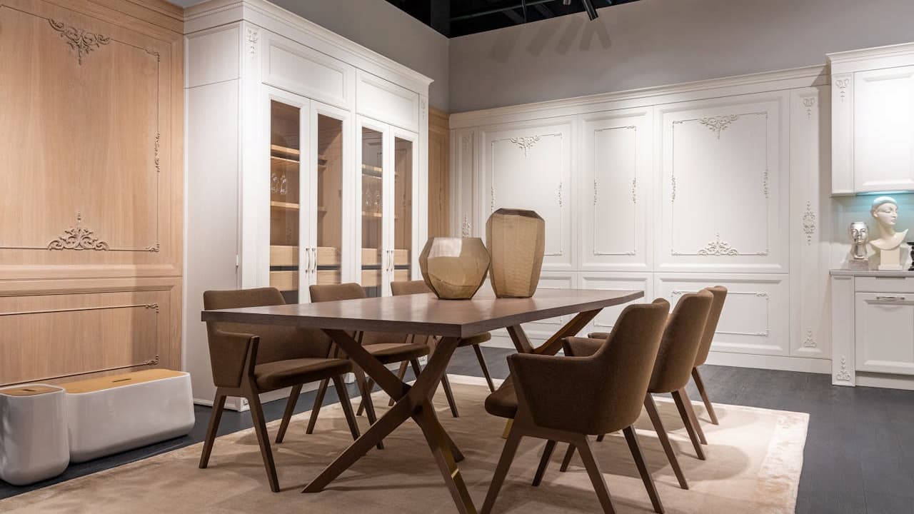 Dining room with a wooden table, six upholstered chairs with angled legs, two decorative vases on the table, large built-in white cabinetry with glass doors and intricate molding, soft beige rug underneath, warm ambient lighting, neutral color palette with wood and white tone