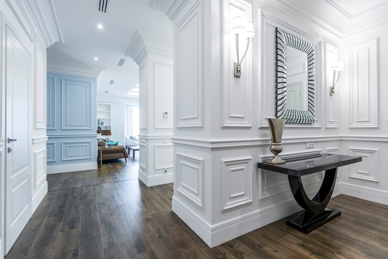 Elegant foyer with white decorative wall moldings, a sleek black console table, silver vase, and a large geometric mirror, leading into a bright living room with light blue accents
