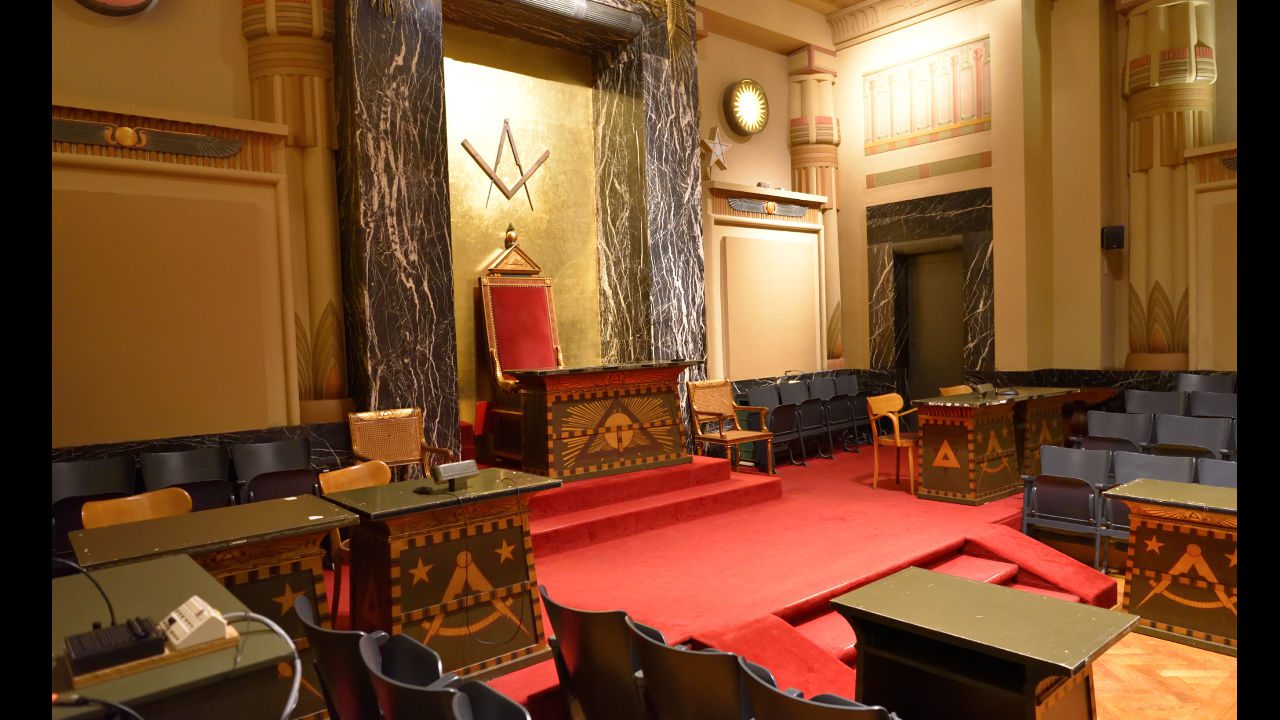 Ornate Freemason lodge room with red carpet, symbolic furniture, and a gold-trimmed central chair beneath Masonic symbols