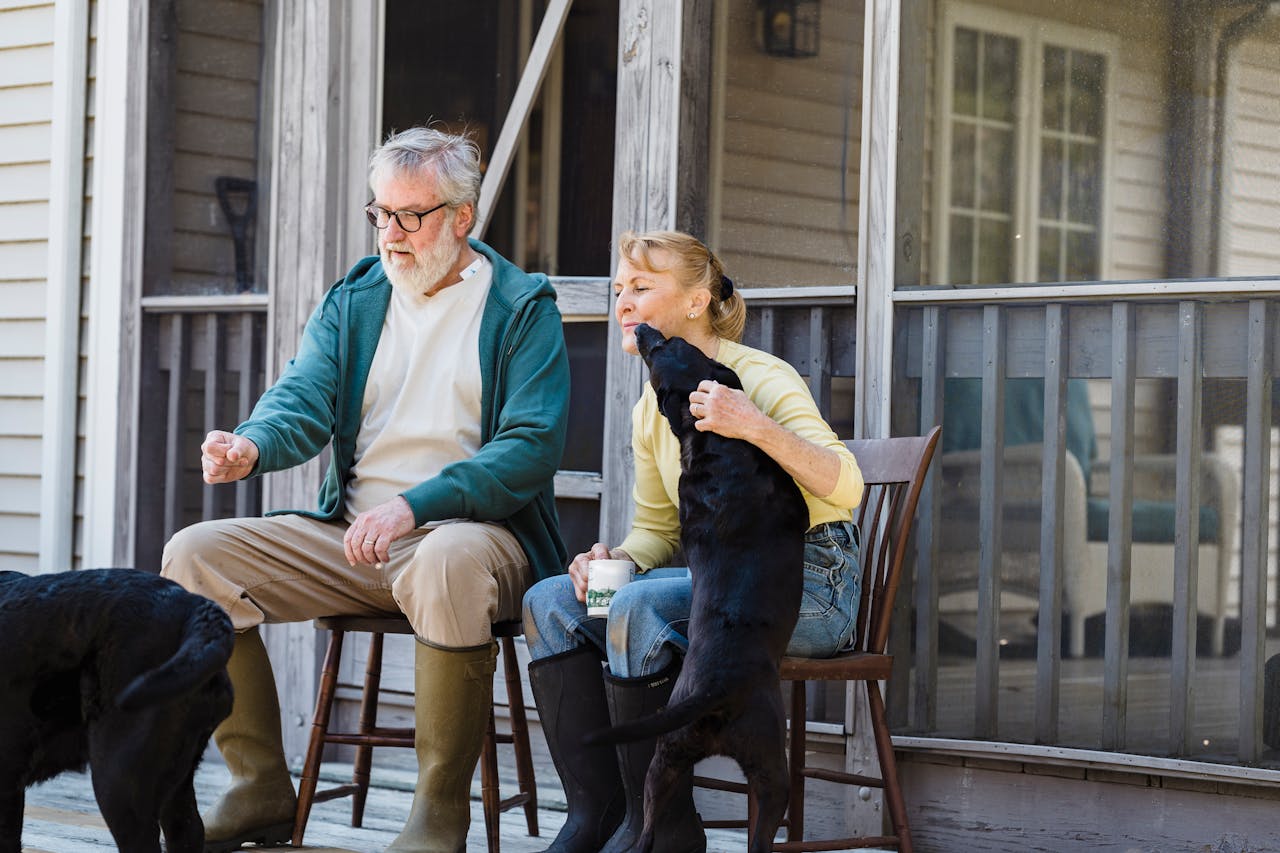 Older couple sitting on wooden chairs on a front porch, interacting with two black dogs, one of which is licking the woman’s face
