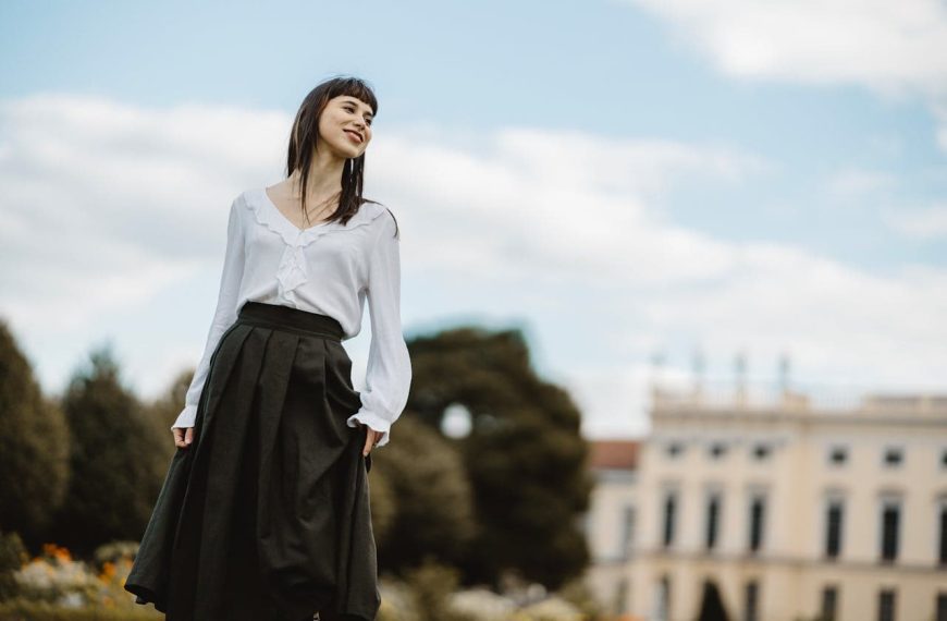 Woman standing outdoors, wearing a white blouse with ruffles and a high-waisted black full-circle skirt, smiling, with a historical building and trees in the background