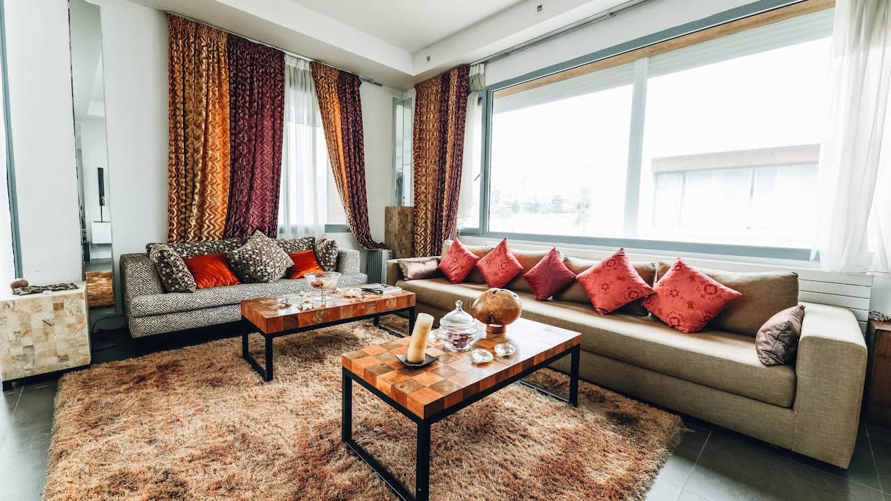 Modern living room, two beige sofas with red and gray cushions, wooden coffee table with decor items, large windows with sheer and heavy curtains