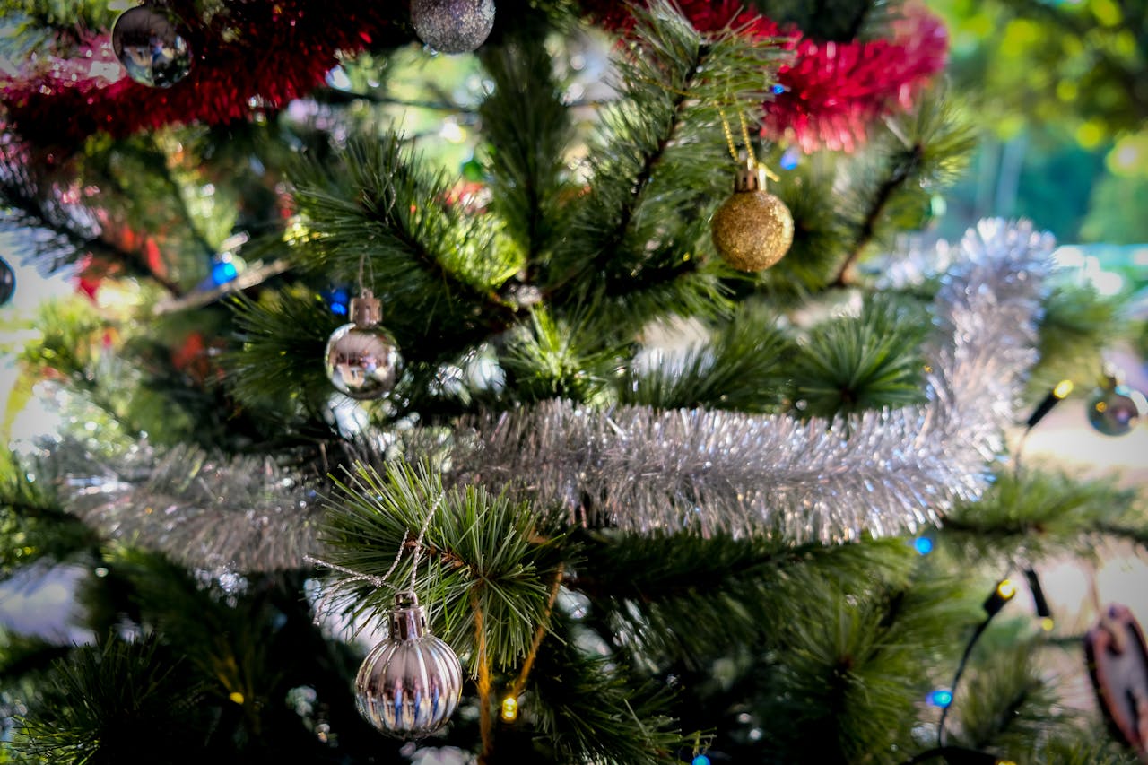 Close-up of a Christmas tree decorated with silver tinsel garland, shiny baubles in silver and gold, and red garland strands, set against a blurred outdoor background