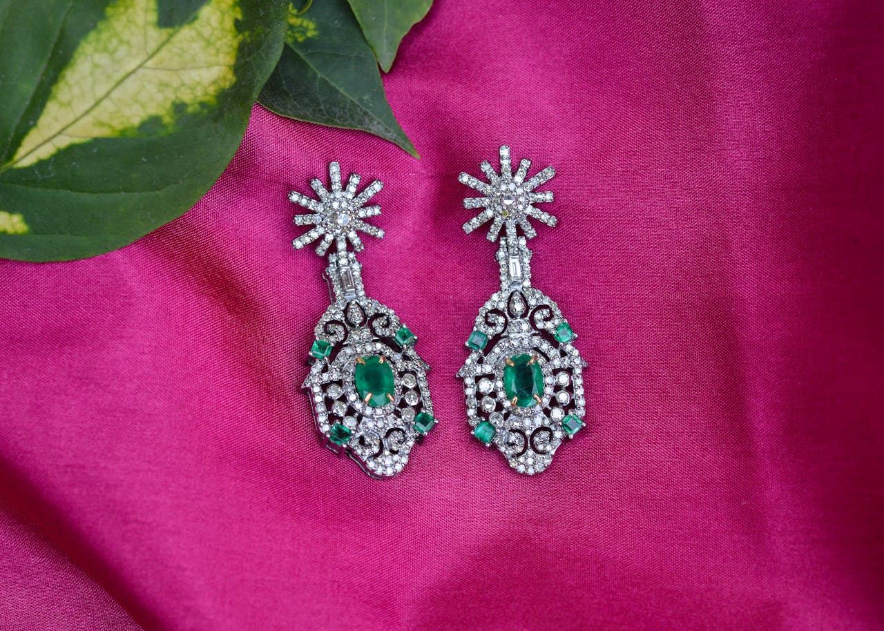 Pair of ornate silver earrings, studded with white stones, featuring a central green gemstone, displayed on a bright pink fabric with green leaves in the corner