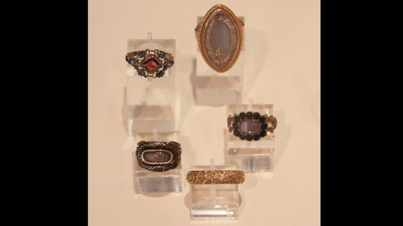 Five antique mourning rings displayed on clear acrylic stands against a beige background, featuring intricate designs with gemstones and engraved details