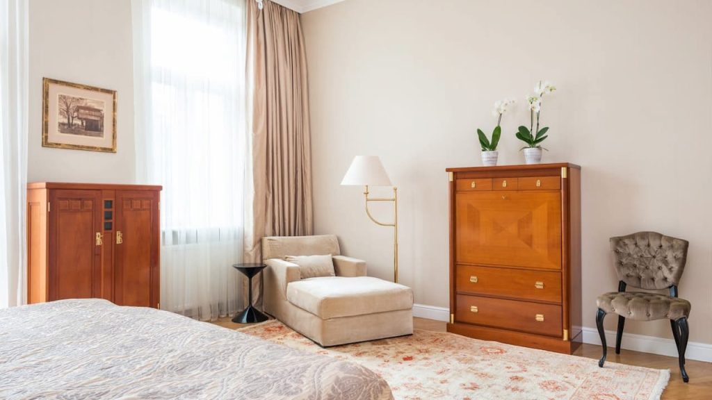 A cozy bedroom corner with a gold floor lamp featuring a distressed gold leaf finish, positioned beside a beige armchair. The room includes light brown curtains, vintage wooden cabinets, a floral rug, and soft lighting from large windows
