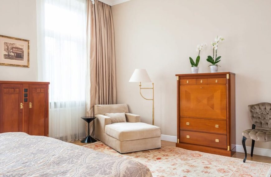 A cozy bedroom corner with a gold floor lamp featuring a distressed gold leaf finish, positioned beside a beige armchair. The room includes light brown curtains, vintage wooden cabinets, a floral rug, and soft lighting from large windows