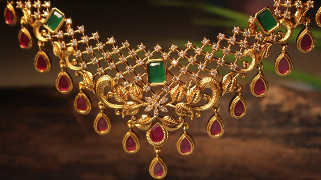 Intricate gold necklace with a lattice design, adorned with green rectangular gemstones, red teardrop-shaped gemstones, and small white stones, displayed against a dark wooden background