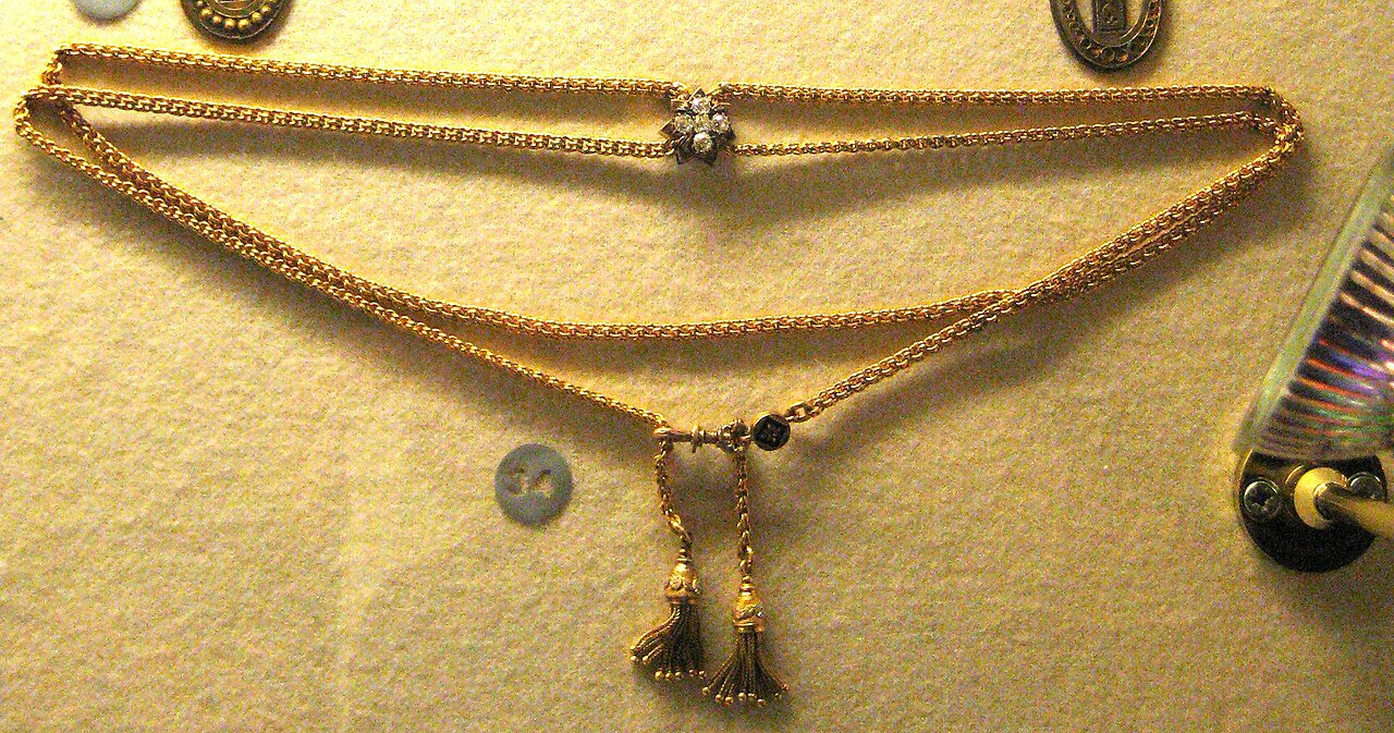 Gold necklace on display, intricate braided design, central pendant with detailed craftsmanship, reflective surface, ambient lighting