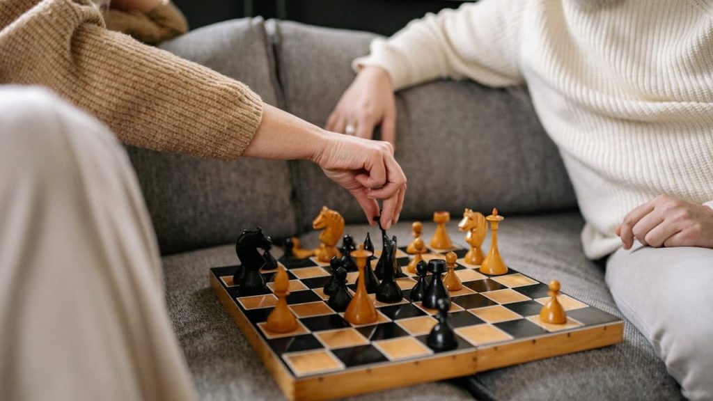Two people sitting on a couch playing chess with a wooden board, one person making a move with a black piece, both dressed in warm sweaters