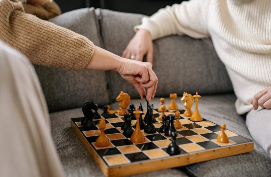 Two people sitting on a couch playing chess with a wooden board, one person making a move with a black piece, both dressed in warm sweaters