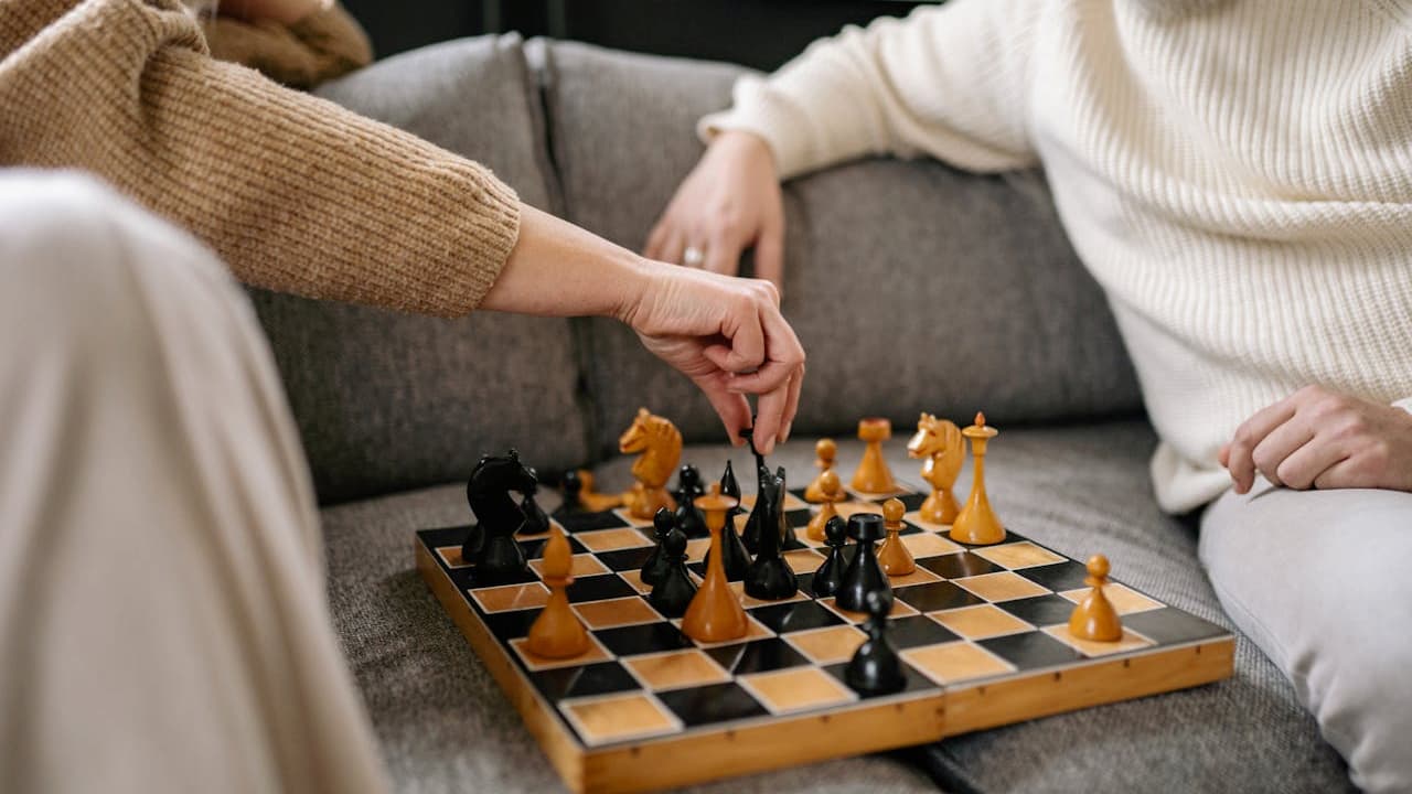 Two people sitting on a couch playing chess with a wooden board, one person making a move with a black piece, both dressed in warm sweaters