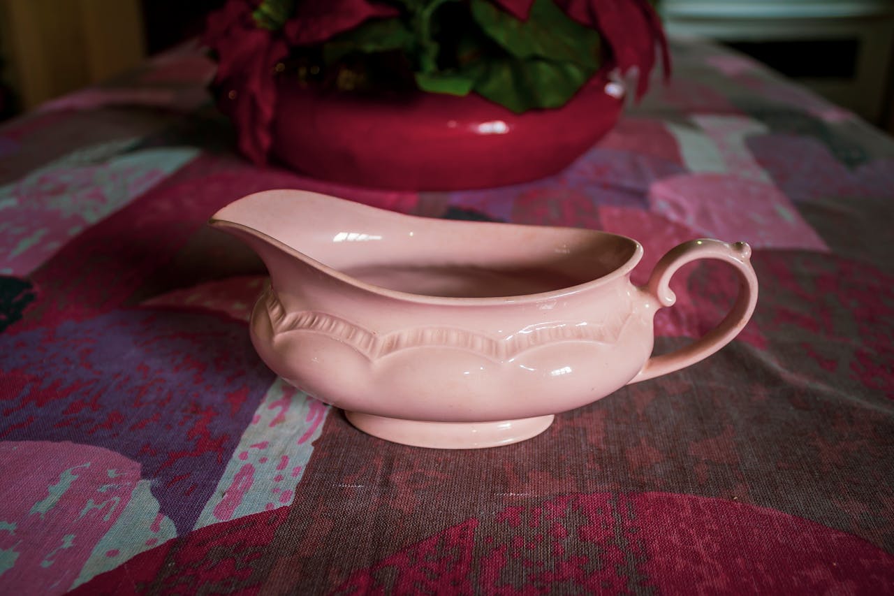 A pale pink ceramic gravy boat or sauce pitcher sits on a patterned tablecloth with burgundy and green tones. In the background, a red planter containing poinsettia flowers adds a festive touch to the scene