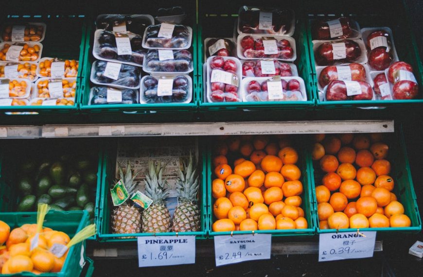 Fresh produce display at a market, assorted fruits and vegetables arranged in green crates, including apples, oranges, lemons, and squash, neatly organized, labeled with prices, natural lighting