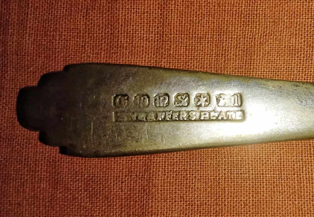 Close-up of a tarnished silver-plated utensil handle, engraved with hallmark-style stamps, marked with the words "SILVER PLATE," displayed on a reddish-brown fabric background