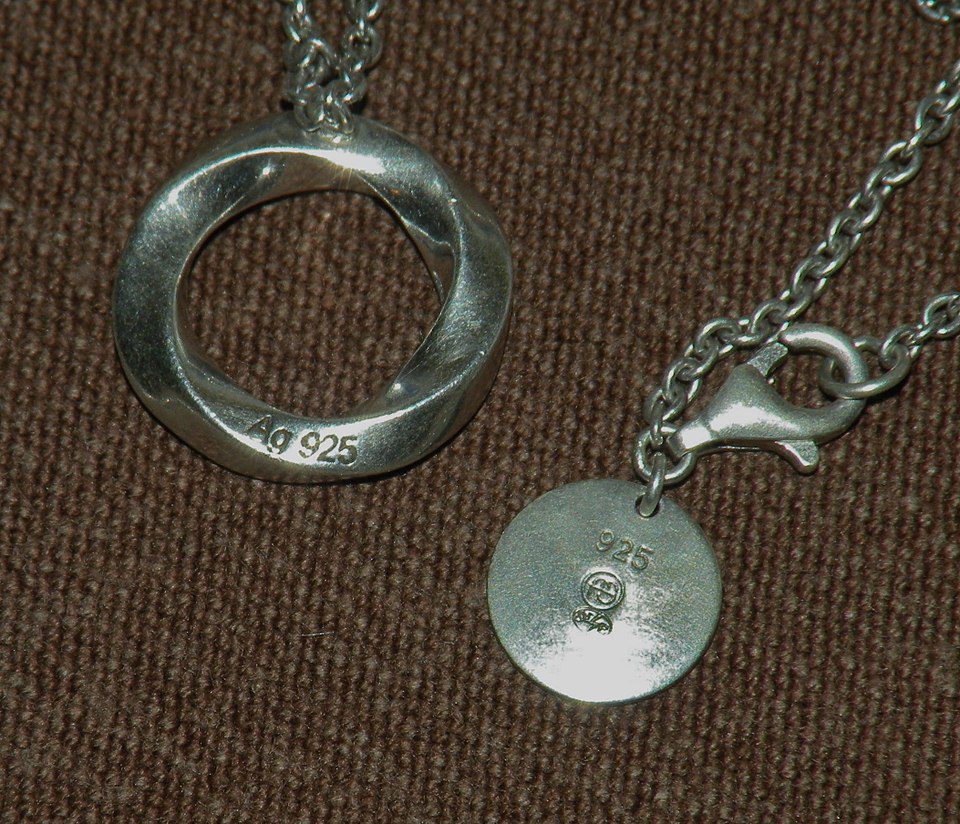 Silver necklace with a circular hollow pendant, round charm with a small gem, lobster clasp, placed on a textured brown surface