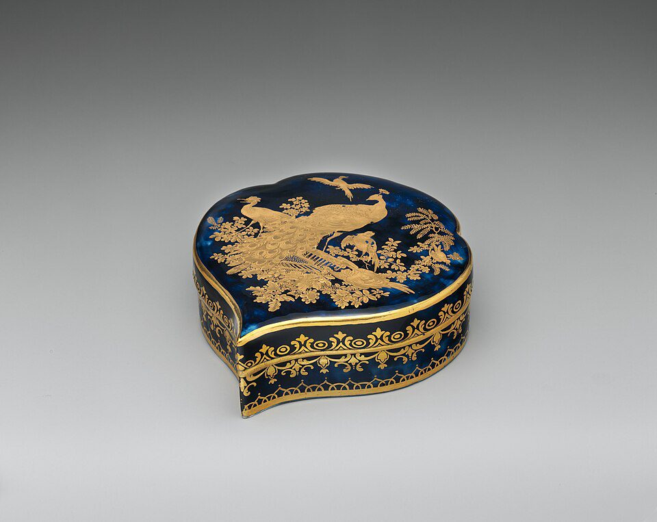 Heart-shaped decorative box, deep blue enamel surface, intricate gold detailing, bird and floral design on lid, ornate gold trim around the sides, antique or vintage style, displayed against a neutral background