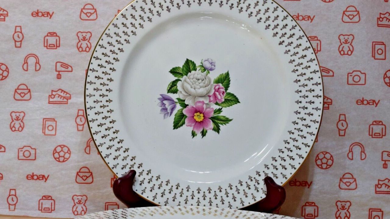 Stack of decorative china plates with a central floral design featuring pink, white, and purple blossoms, surrounded by a gold-trimmed border with repeating black floral patterns