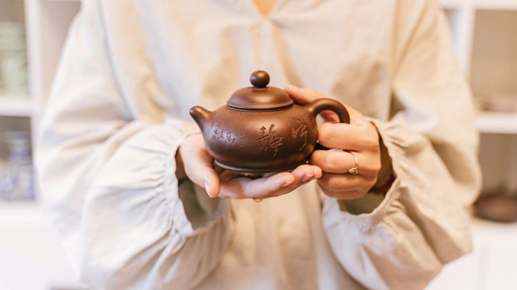 Person in a light-colored robe holding a small brown teapot with engraved Chinese characters, displayed with both hands