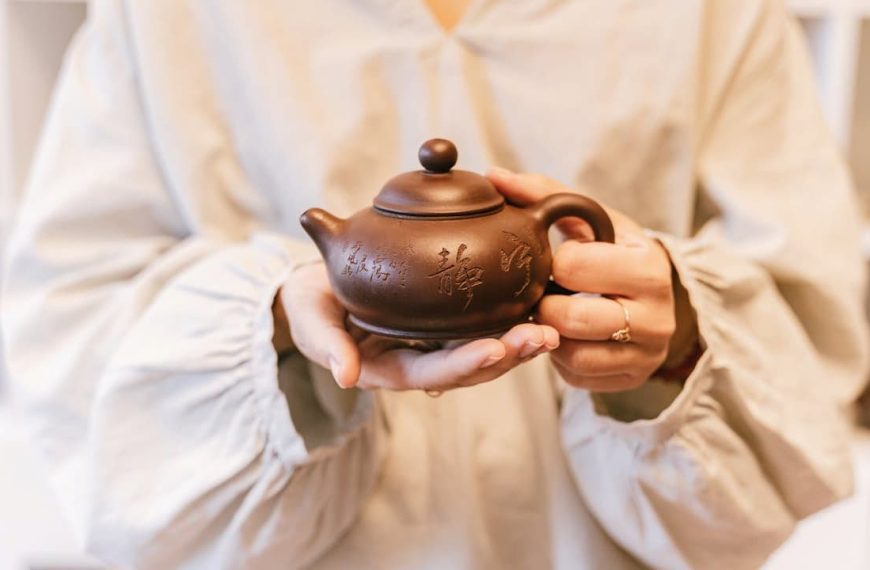 Person in a light-colored robe holding a small brown teapot with engraved Chinese characters, displayed with both hands