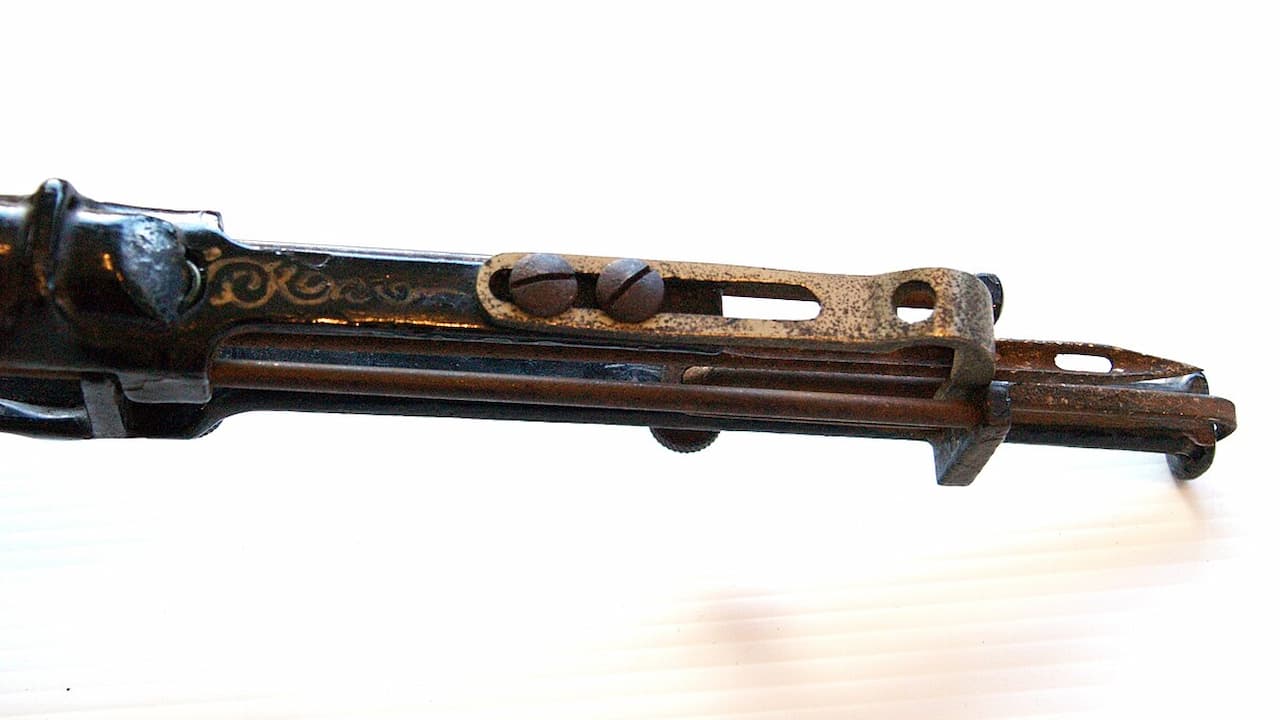 Close-up of an antique firearm or tool with visible scratches, rust, and surface imperfections, showing decorative etching and aging on the metal parts