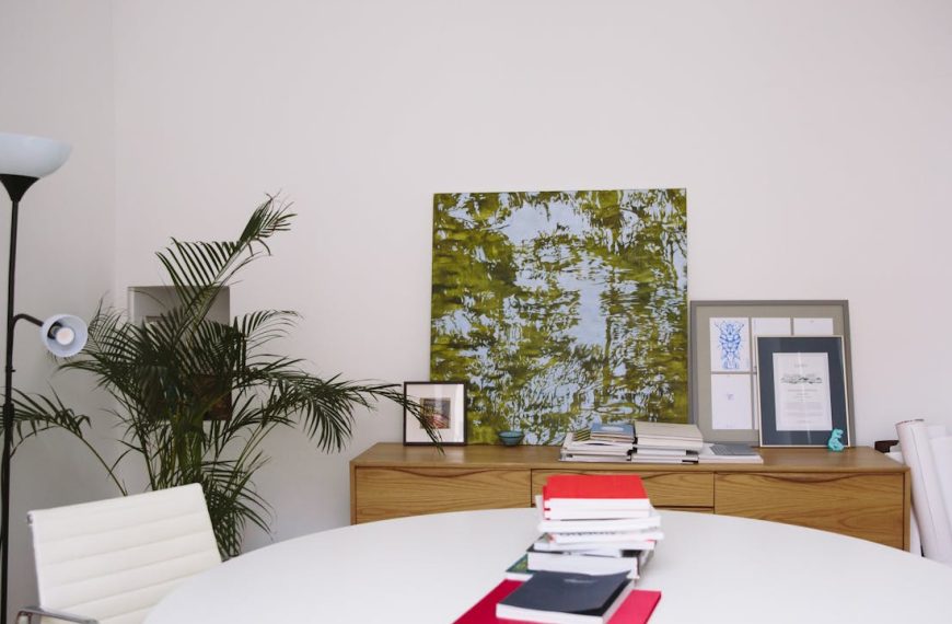 Round white table with documents and a laptop, wooden sideboard against a plain wall, abstract green artwork above, framed pictures arranged on the sideboard, tall indoor plant in the corner, modern floor lamp nearby, clean and minimal home office or study area