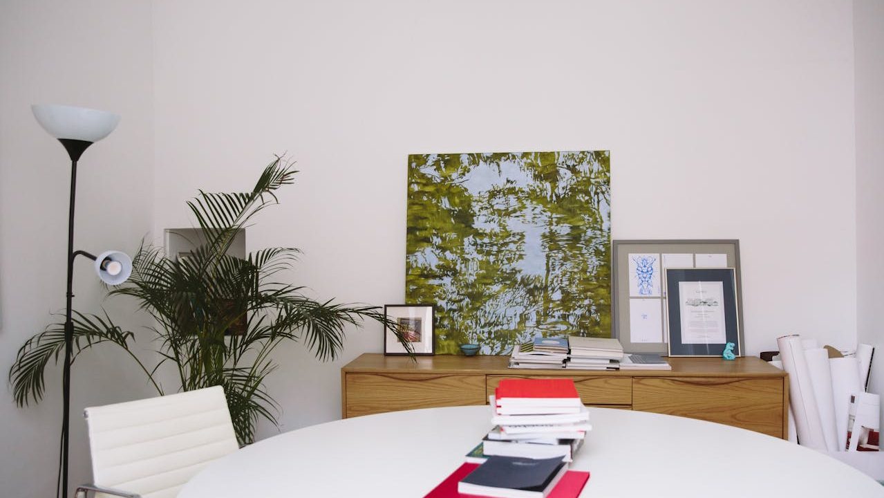 Round white table with documents and a laptop, wooden sideboard against a plain wall, abstract green artwork above, framed pictures arranged on the sideboard, tall indoor plant in the corner, modern floor lamp nearby, clean and minimal home office or study area