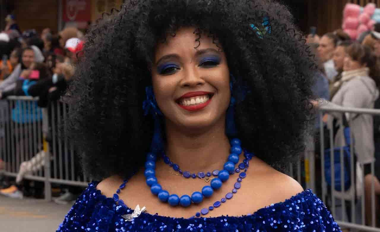 Woman with voluminous curly hair, wearing a sparkling blue sequin outfit, bold blue eye makeup, layered blue beaded necklaces, and earrings, smiling in front of a crowd behind metal barricades