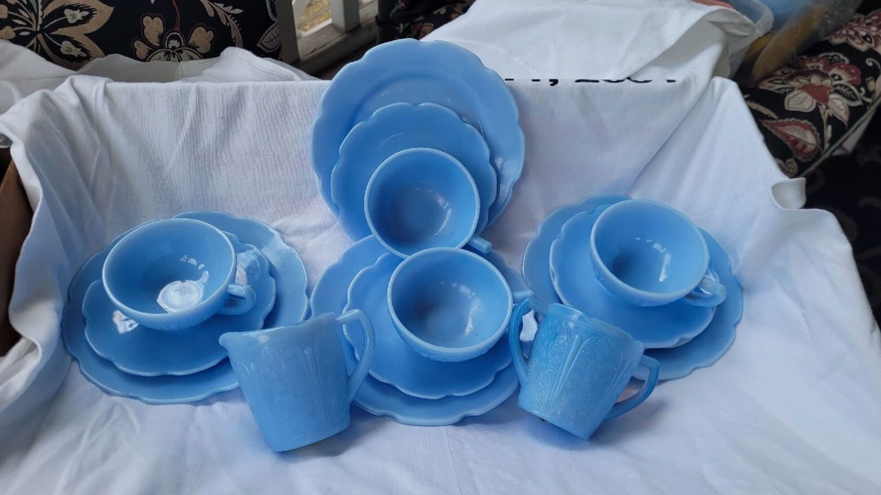 Vintage Jeanette Delphite blue glassware set, including cups, saucers, plates, and mugs, arranged on white fabric