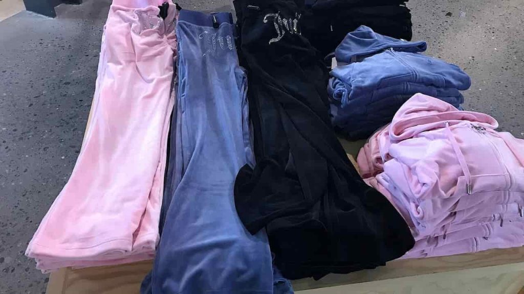 Folded velour Juicy Couture tracksuits in pink, blue, and black, neatly displayed on a wooden table, inside a clothing store with a concrete floor