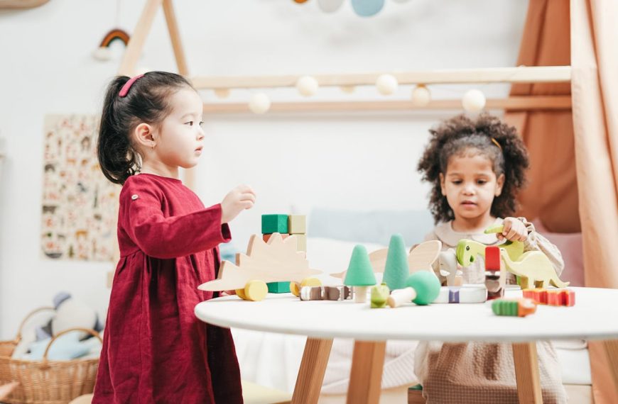 Two young girls, playing with colorful wooden toys, at a round white table, in a bright and cozy playroom, with shelves and decor in the background
