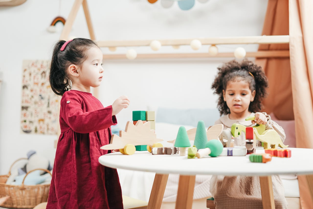 Two young girls, playing with colorful wooden toys, at a round white table, in a bright and cozy playroom, with shelves and decor in the background