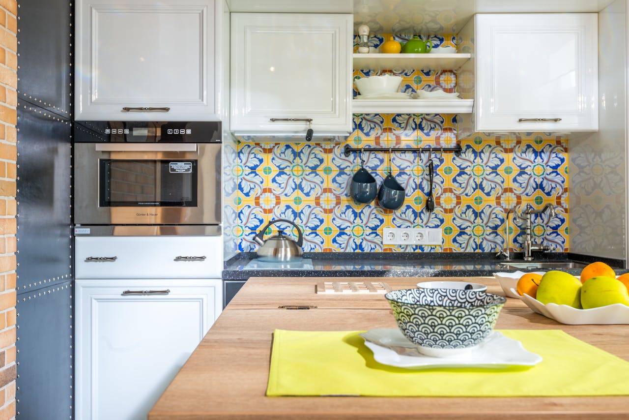 Kitchen interior, colorful tiled backsplash, white cabinets, built-in microwave, stovetop with hanging utensils above, wooden countertop with yellow placemat