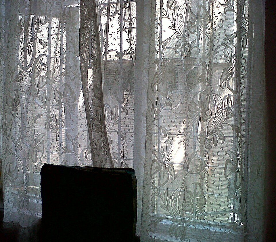 Sunlight filtering through sheer, white lace curtains covering a window, with tree branches visible outside