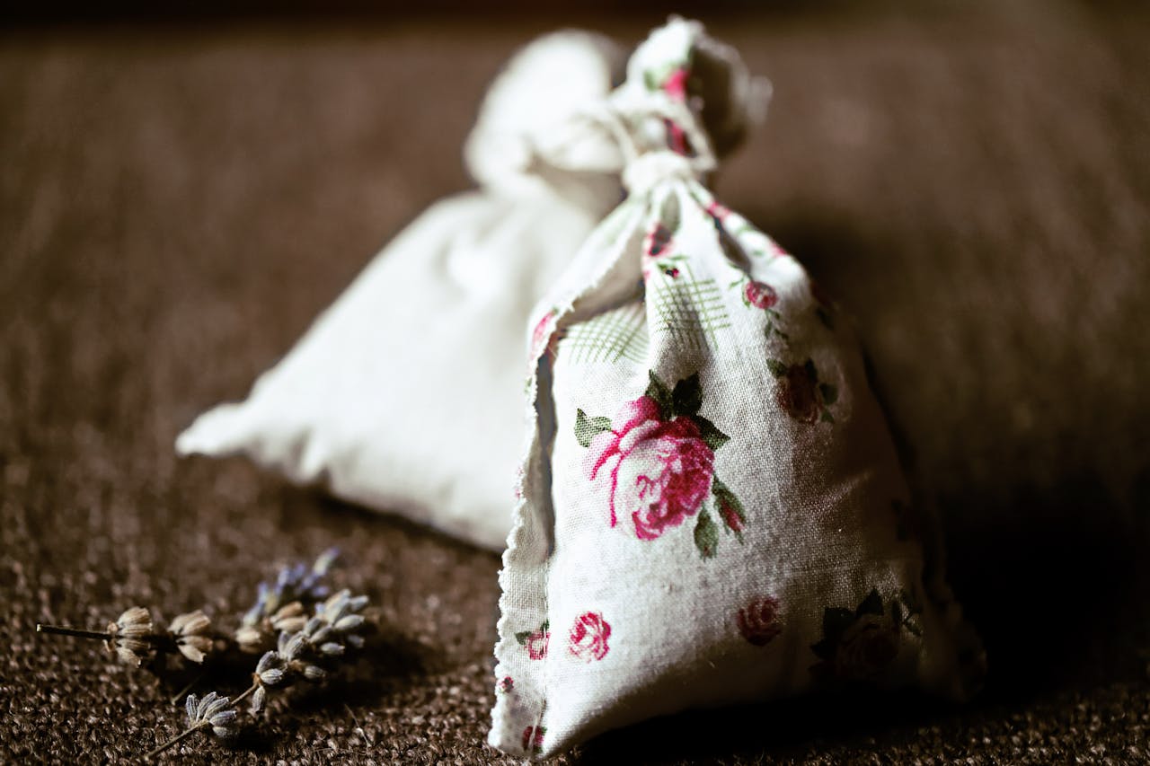 A small fabric sachet with pink rose pattern tied at the top, resting on a dark brown surface. A dried lavender sprig lies beside it, suggesting it's an aromatic potpourri or herbal sachet