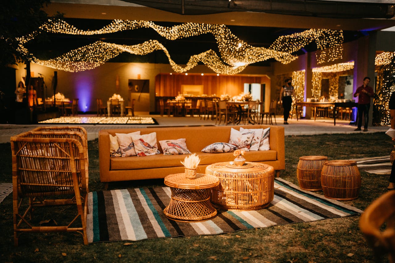 Cozy wicker furniture on a striped rug, warm string lights draped above, decorative cushions on a tan sofa, festive ambiance, tables set for dining in the background, soft ambient lighting, relaxed and elegant atmosphere
