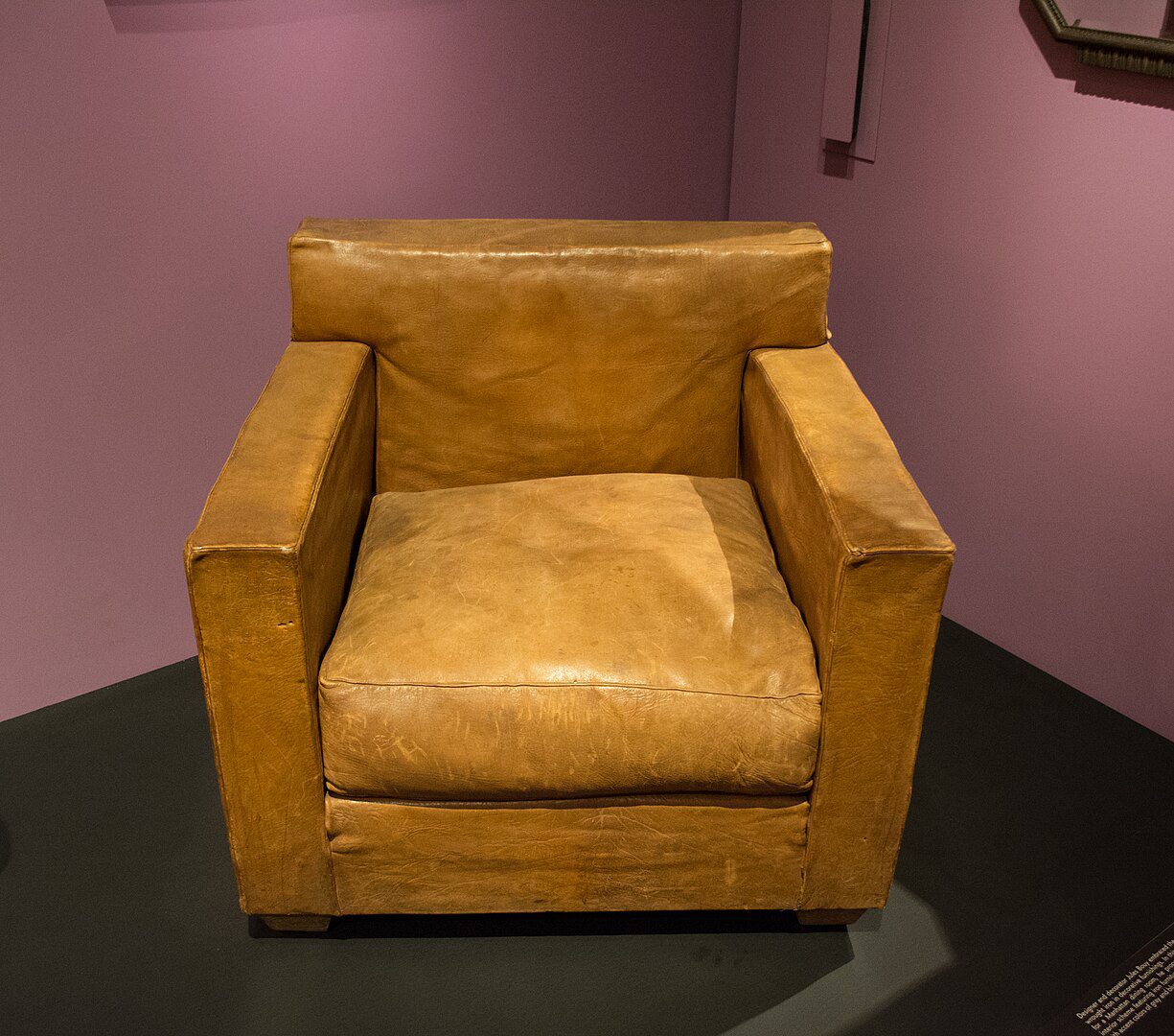 Brown leather armchair, wide square armrests, deep cushioned seat, boxy design, set against a purple wall, placed on a dark floor platform, soft worn texture, museum or showroom setting, modern minimalist style