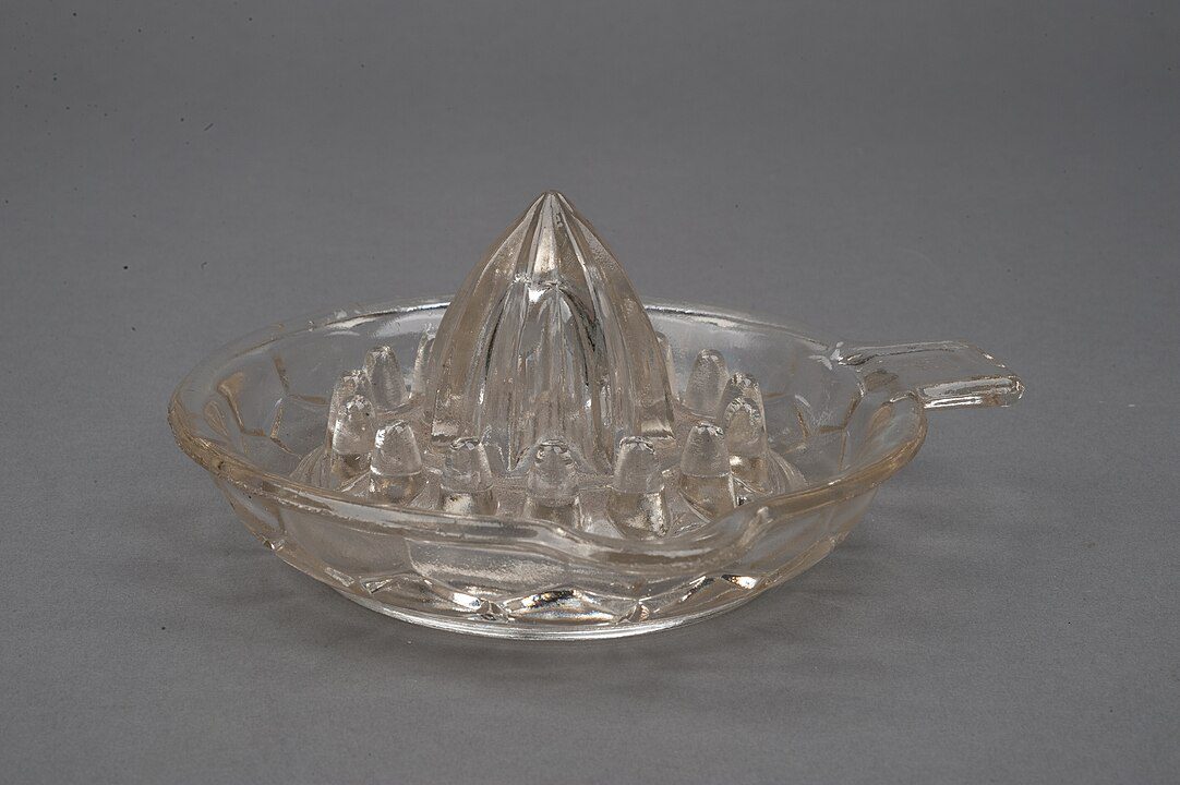 Clear glass citrus juicer, conical reamer center, ridged juice-catching base, small handle on side, set against plain gray background, vintage kitchen tool design