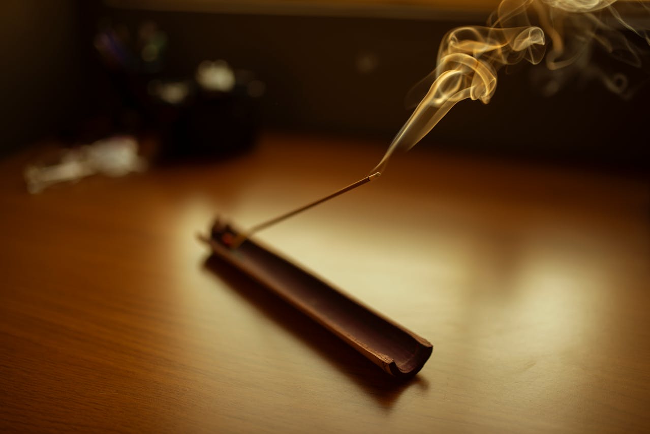 Burning incense stick, placed in a wooden holder, thin trail of smoke rising, set on a wooden surface, warm ambient lighting, calm and relaxing atmosphere