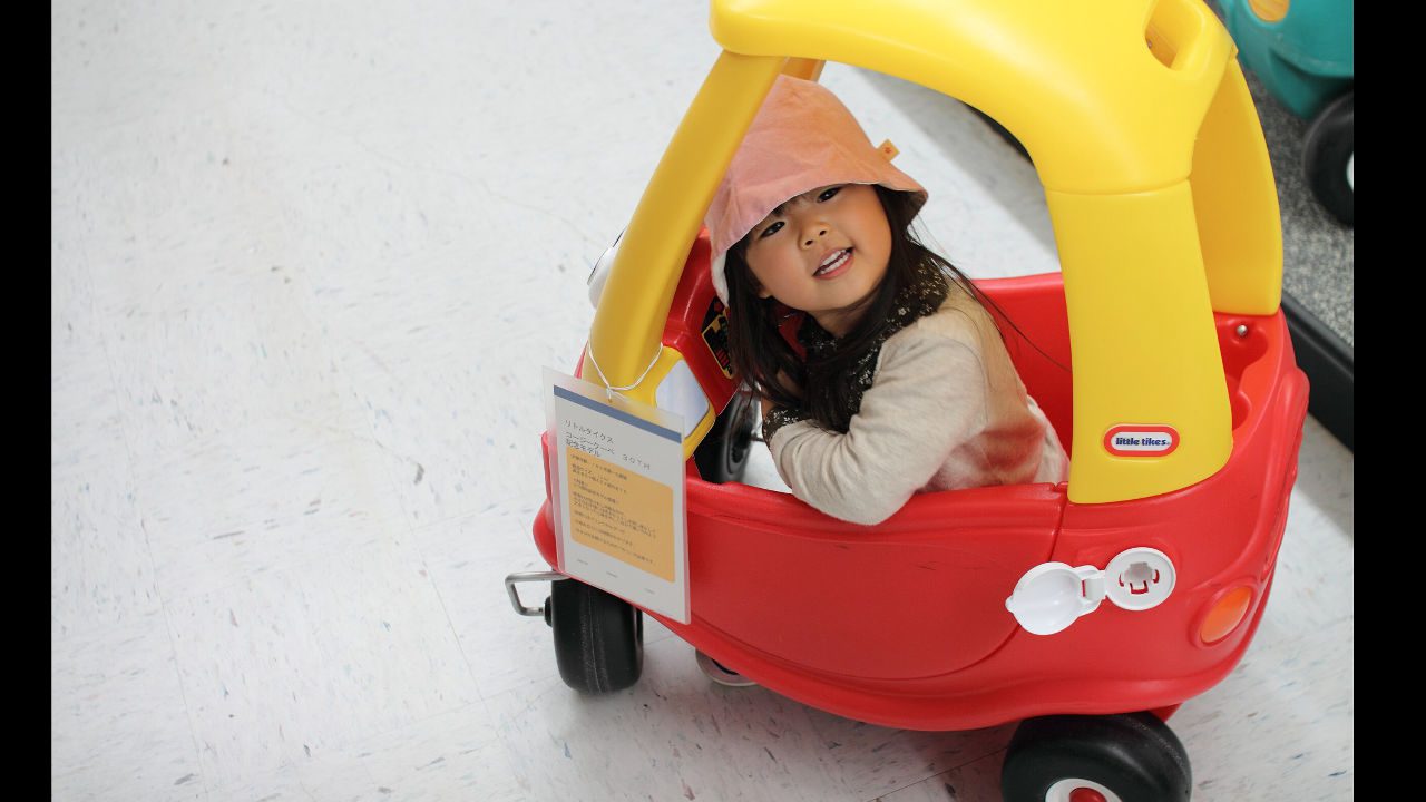 Child wearing a pink hat sits inside a red and yellow Little Tikes Cozy Coupe car on a tiled floor indoors