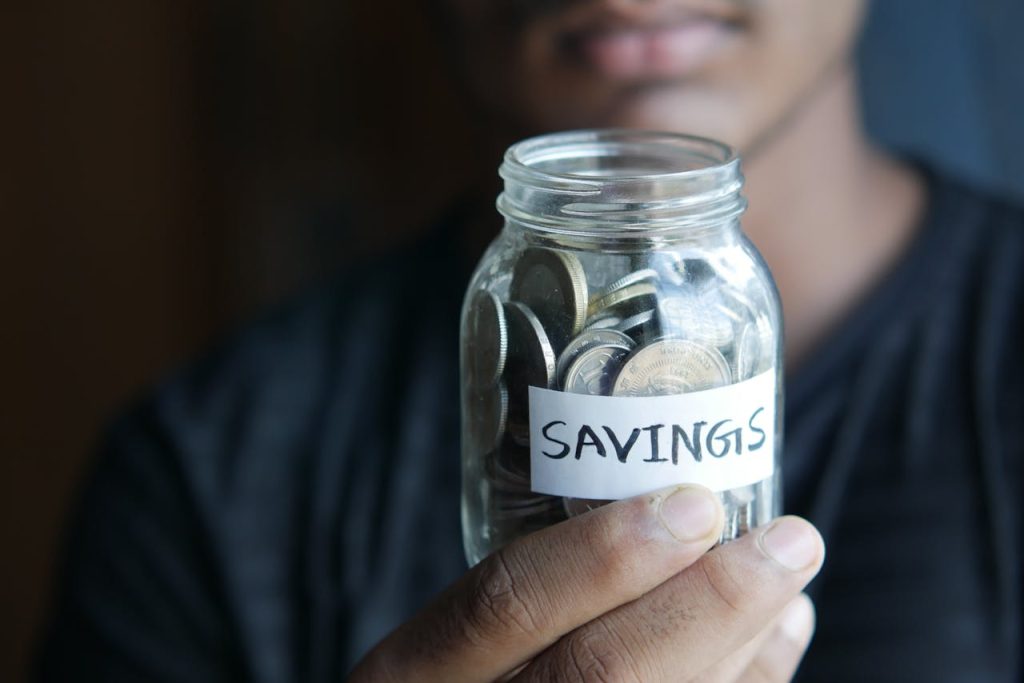 Person holding a glass jar labeled "savings", filled with cash and coins, close-up view, dimly lit background, dark clothing, focus on financial planning