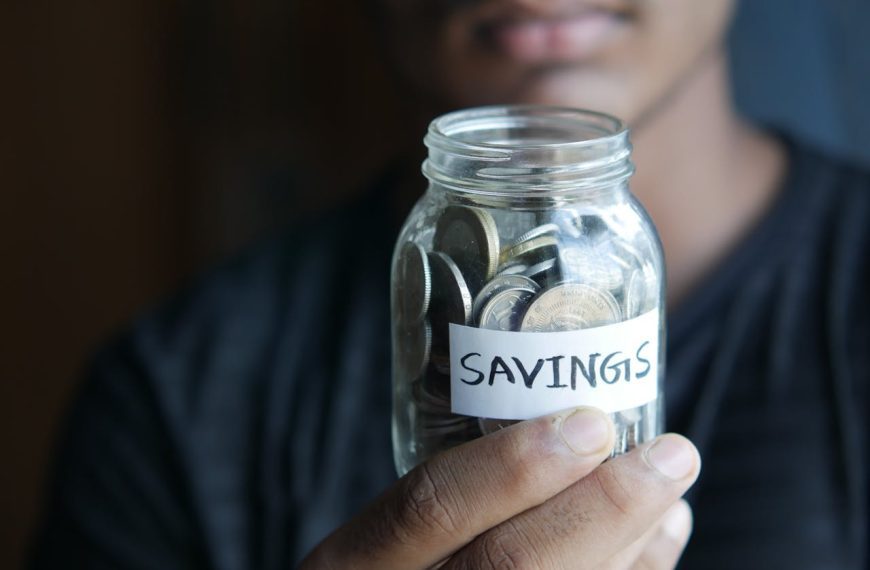 Person holding a glass jar labeled "savings", filled with cash and coins, close-up view, dimly lit background, dark clothing, focus on financial planning