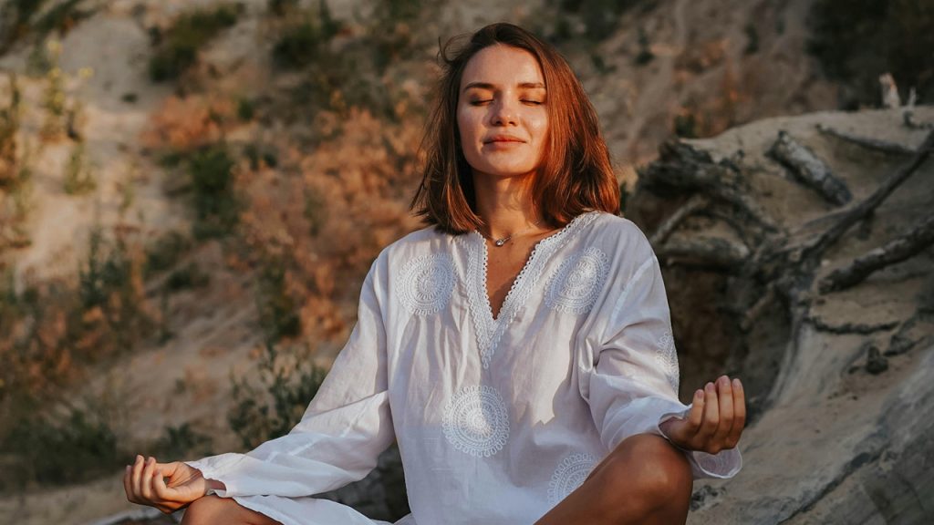Meditation relaxing, Woman meditating outdoors, eyes closed, wearing white top, sitting calmly in natural setting, sunset light