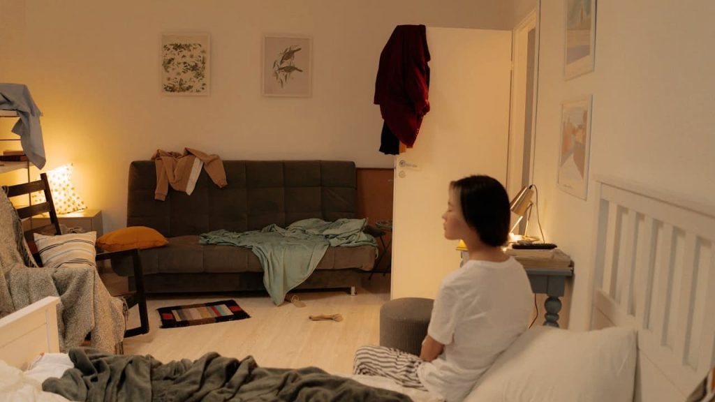 Young woman sitting on the edge of a messy bed in a dimly lit bedroom, with a cluttered sofa, chairs with draped clothes, a small table with a lamp, and open door with clothes hanging on it