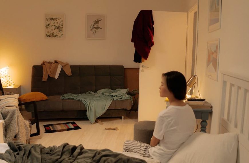 Young woman sitting on the edge of a messy bed in a dimly lit bedroom, with a cluttered sofa, chairs with draped clothes, a small table with a lamp, and open door with clothes hanging on it