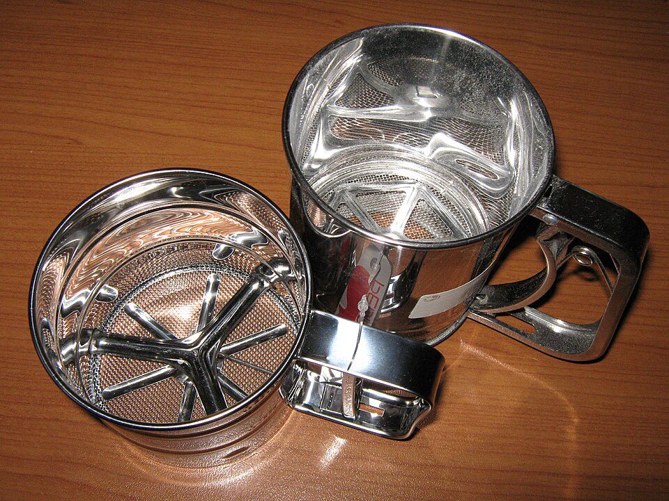 Metal flour sifter, hand-crank handle, stainless steel body, mesh screen inside, rotary blades for sifting, used for baking, placed on wooden surface, two sifters shown