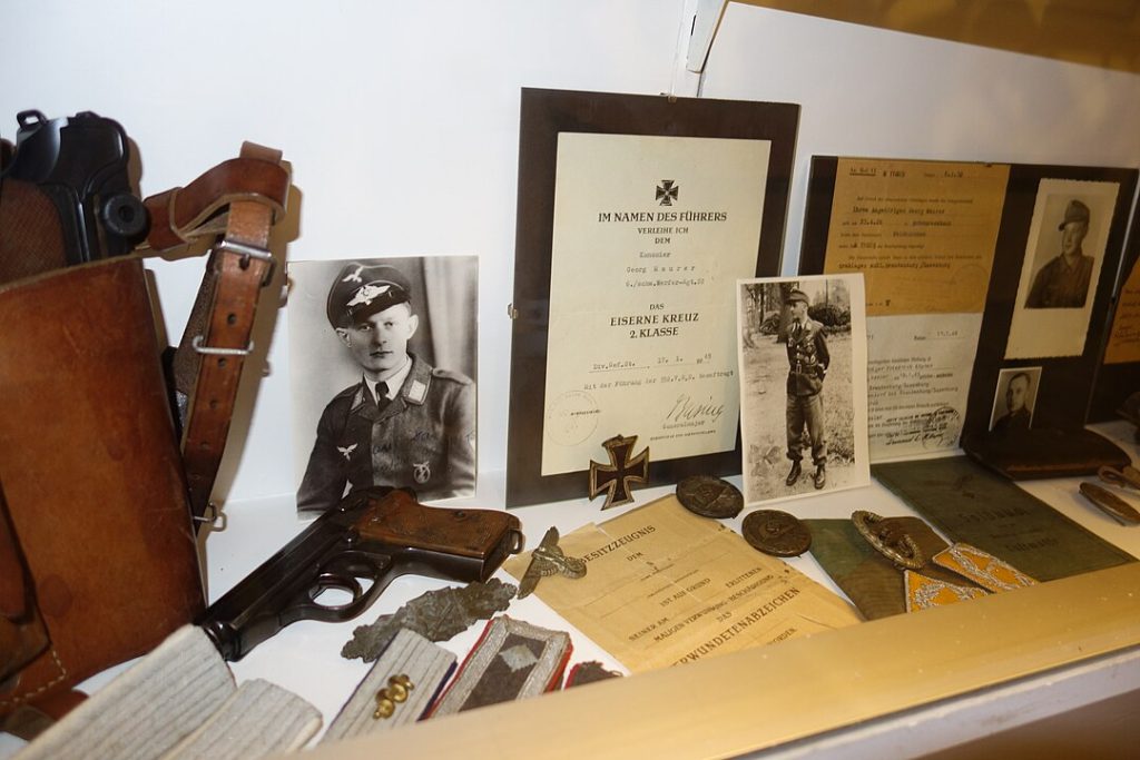 Vintage military memorabilia display, black-and-white soldier portraits, framed certificate, leather holster, service medals, old documents, and keys arranged on a table