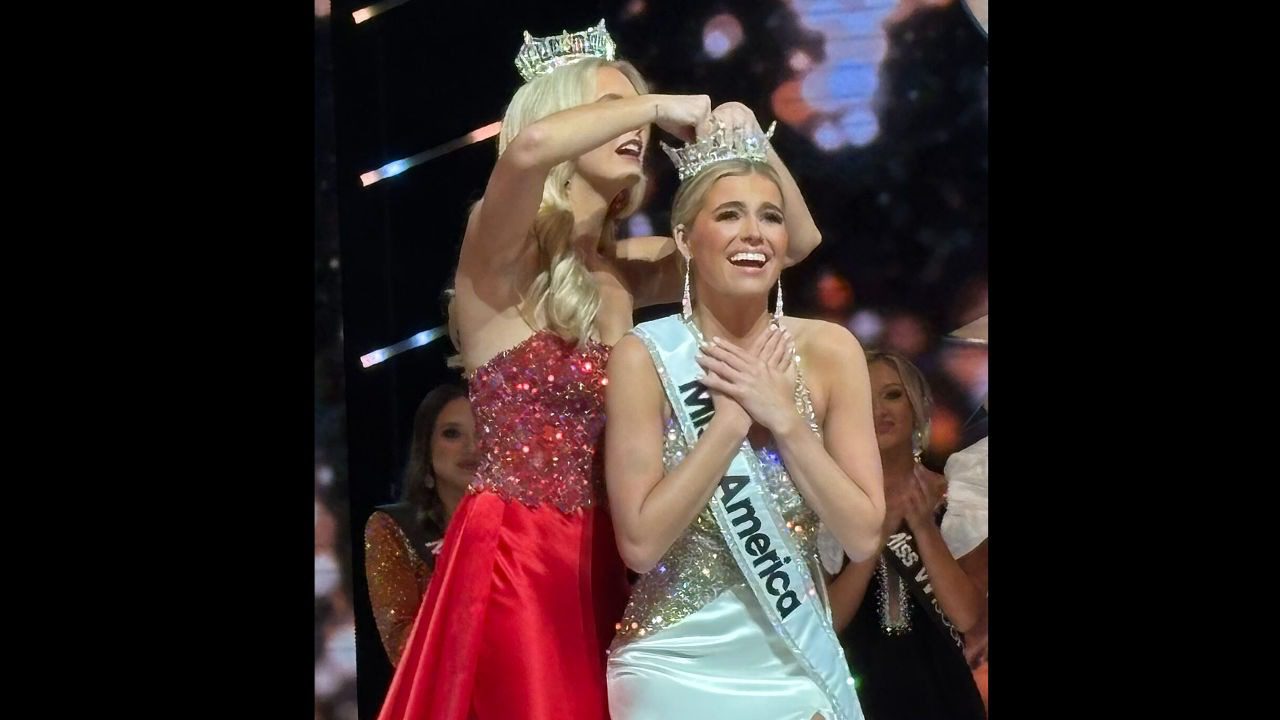 Smiling woman in white gown crowned as Miss America on stage, surrounded by contestants in formal dresses