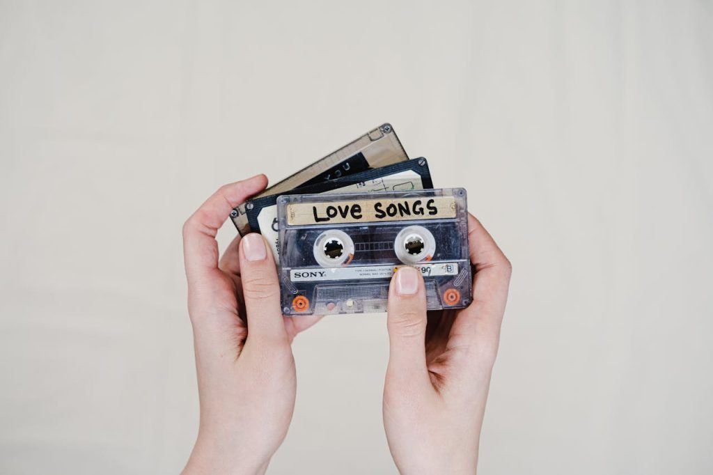 Hands holding three vintage cassette tapes against a plain light background. The front cassette has a handwritten label reading "LOVE SONGS" and is a Sony brand tape