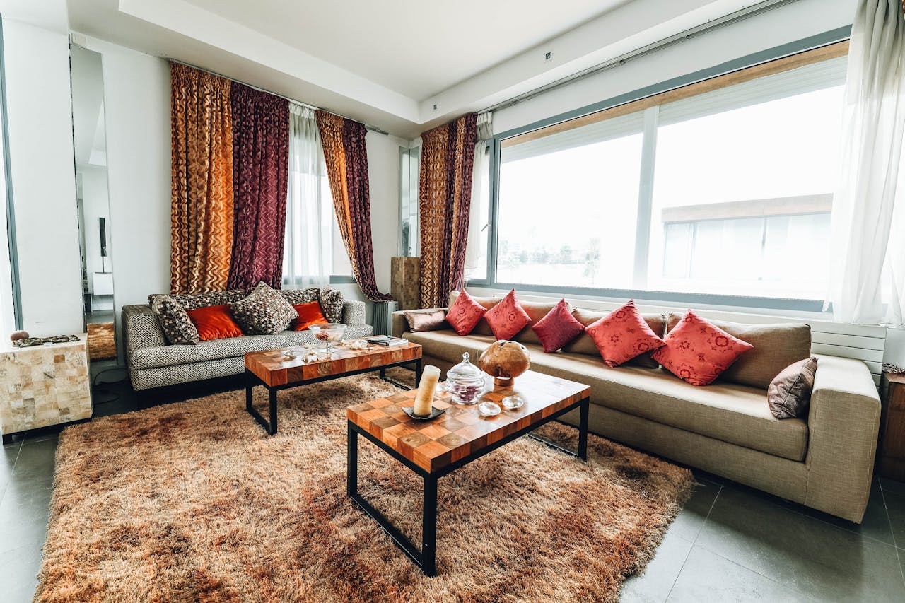 Modern living room, large windows with natural light, two beige sofas with red cushions, wooden coffee tables, plush brown rug, decorative curtains in gold and maroon, cozy and stylish interior